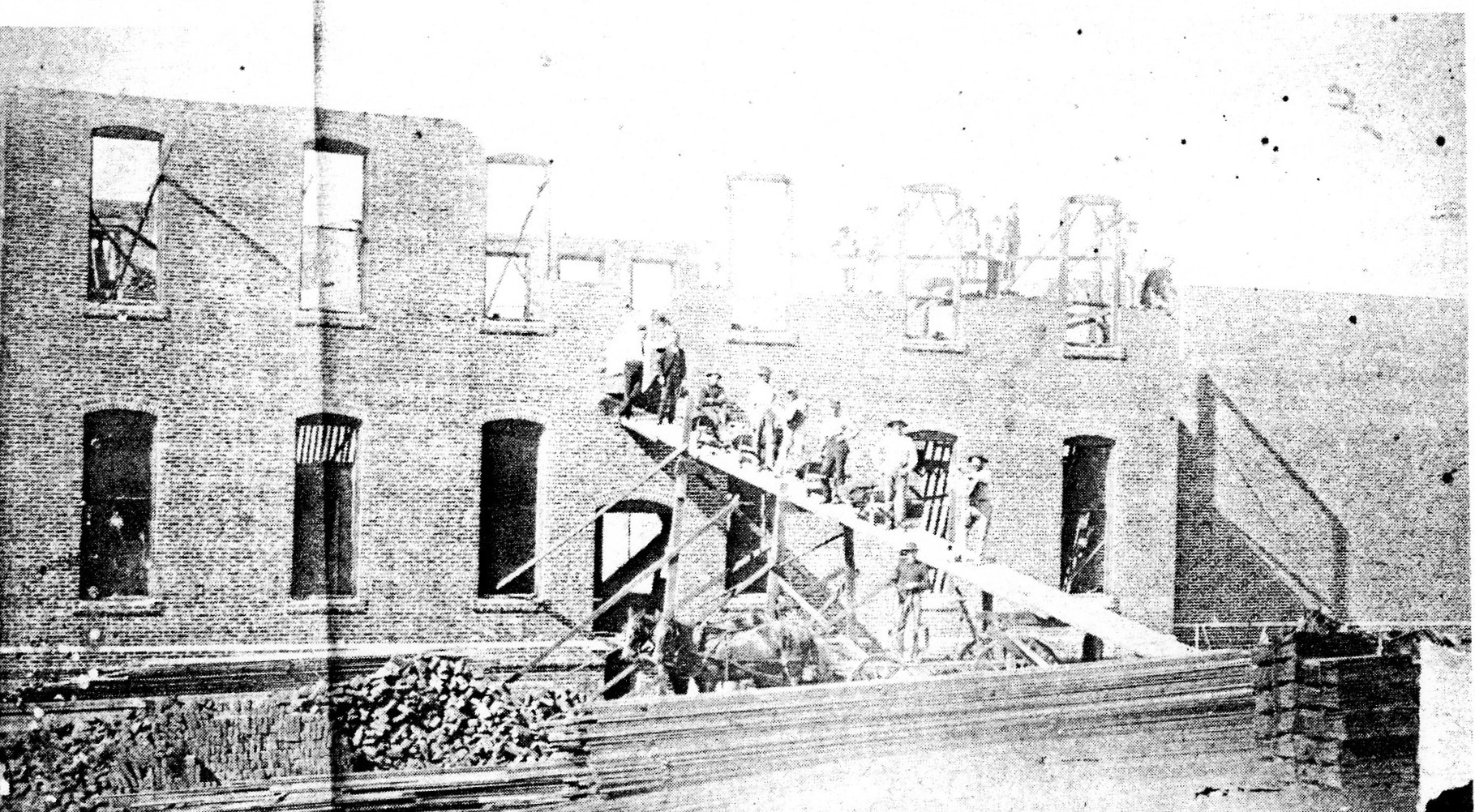 Historic black-and-white photograph of workers on a temporary ramp and scaffolding outside a brick building under construction.