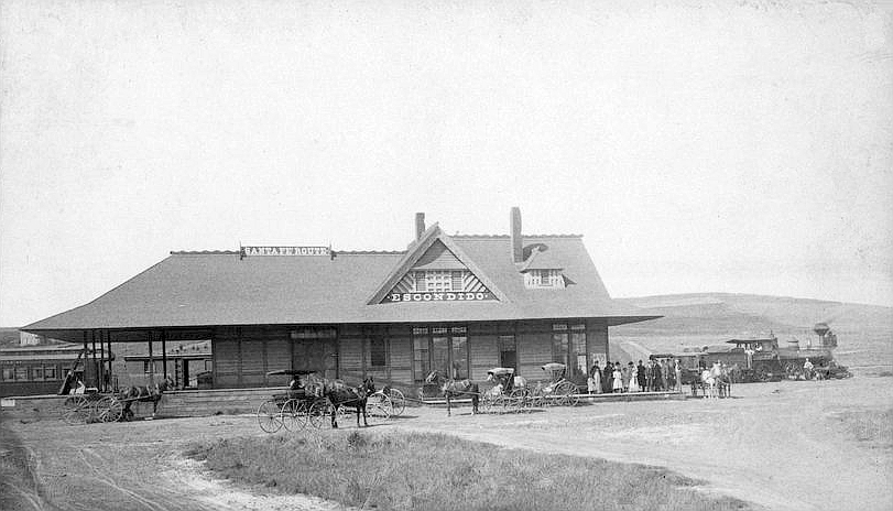 Black-and-white side view of Escondido’s wooden Santa Fe depot beside railroad tracks, with people and rail cars nearby.