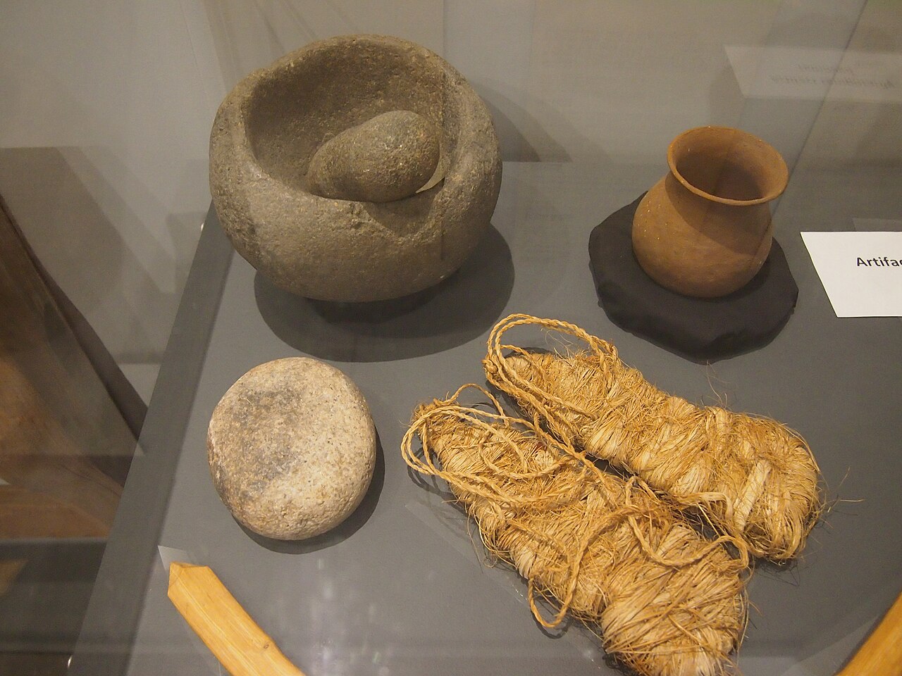 Stone mortar and pestle, ceramic vessel, and fiber materials arranged in a museum display.
