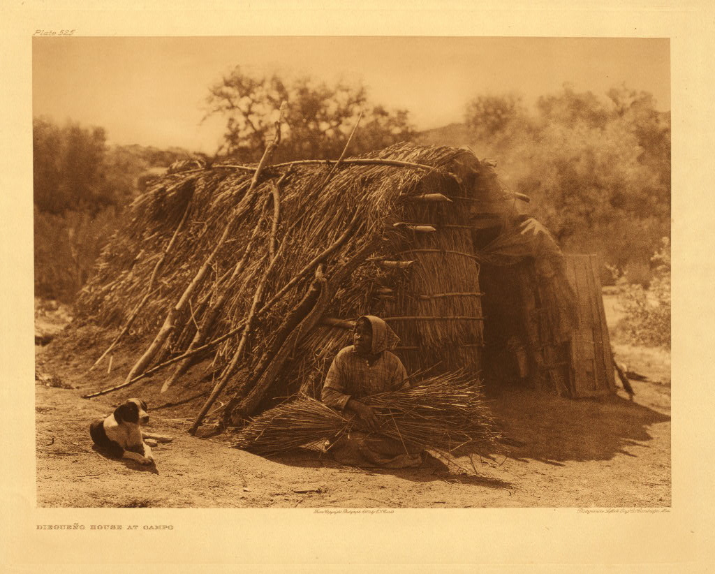 Sepia-toned photograph of a Kumeyaay woman seated with gathered plant material beside a brush house at Campo, with a dog nearby.