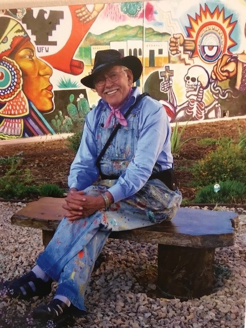 Portrait of muralist Salvador R. Torres seated on a bench with a colorful Chicano Park mural behind him.