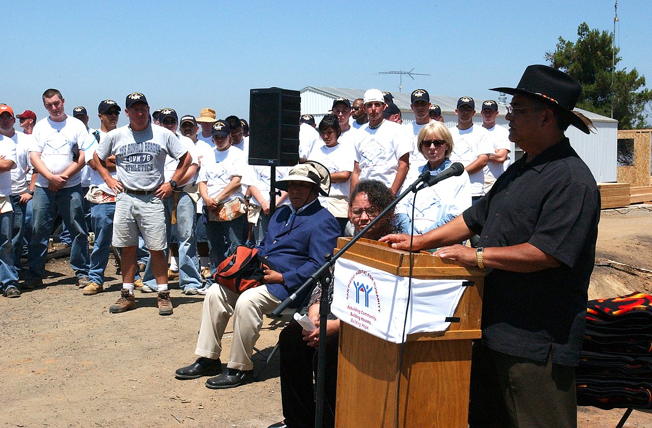 Allen Lawson speaks at a podium during a 2004 community home-building event while participants stand behind him.